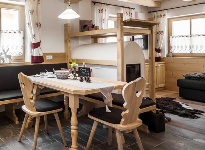Ein gedeckter Holztisch mit zwei Holzstühlen, ein rustikaler Ofen mit Eckbank in einem gemütlichen, holzgetäfelten Zimmer