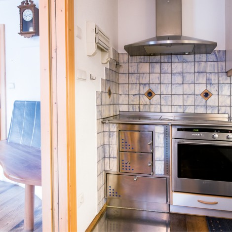 Kleine Küche mit Edelstahlherd, Backofen, Dunstabzugshaube und gefliester Wand neben einem offenen Durchgang zu einem Raum mit Holztisch und Tür