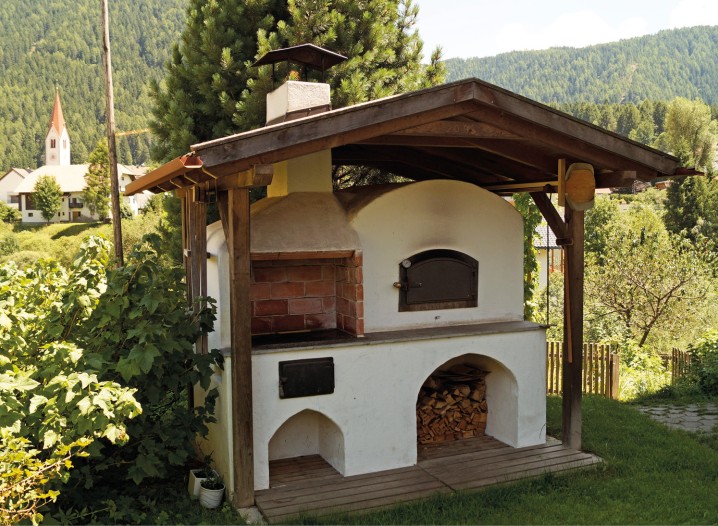 Freistehender, weiß verputzter Holzbackofen mit Schornstein und Holzlager unter einem Holzdach in einer grünen Berglandschaft