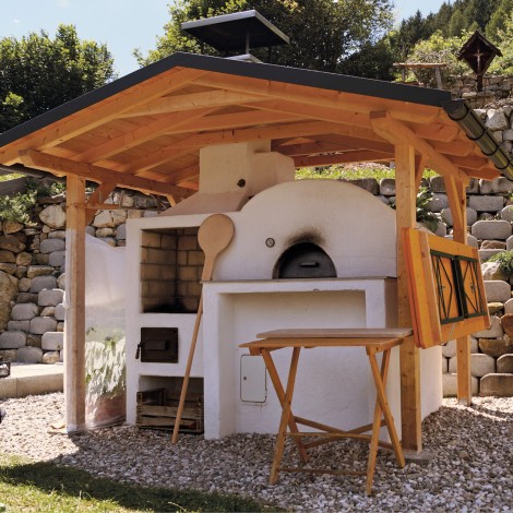 Freistehender Holzbackofen mit Dach, Holzlöffel und Klapptisch in einem Garten mit Steinmauer im Hintergrund