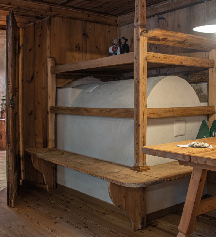 Holzstube mit eingebautem Ofenbankbett, Holztisch mit Bank und Topfpflanze, offene Tür zum Flur