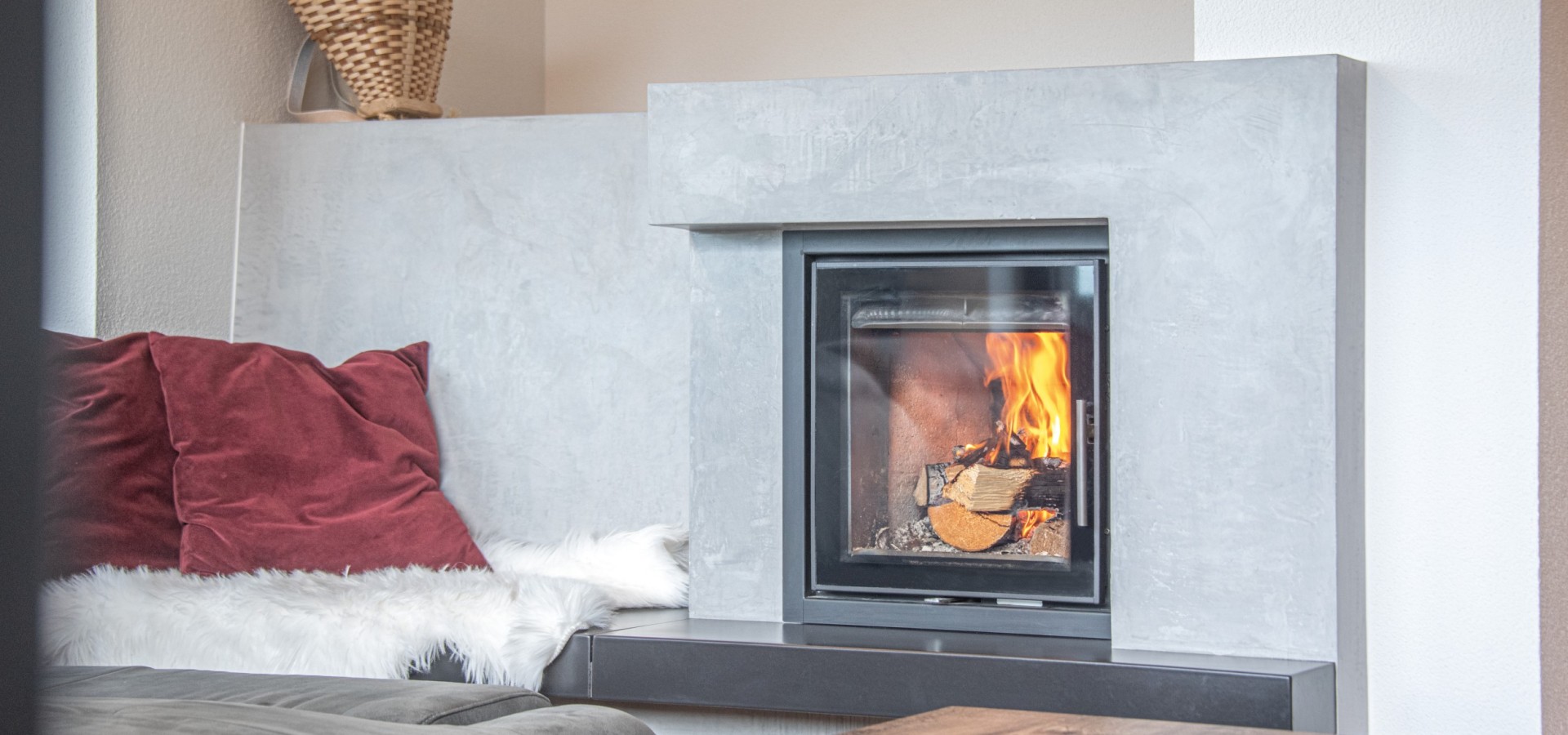 Moderner Kamin mit brennendem Holz in einem hellen Wohnzimmer neben einem Sofa mit rotem Kissen und weißem Fellüberwurf