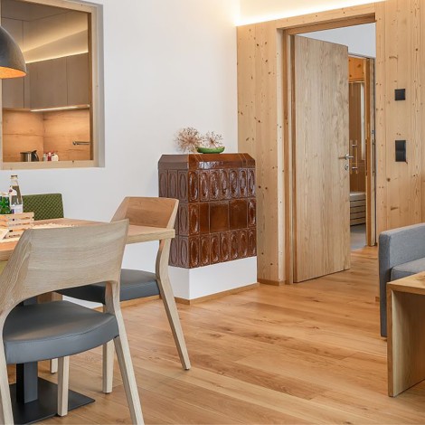 Essbereich mit Holztisch, Stühlen und grün gepolsterter Bank neben einem braunen Kachelofen in einem Raum mit Holzboden und offener Tür zu einem weiteren Zimmer