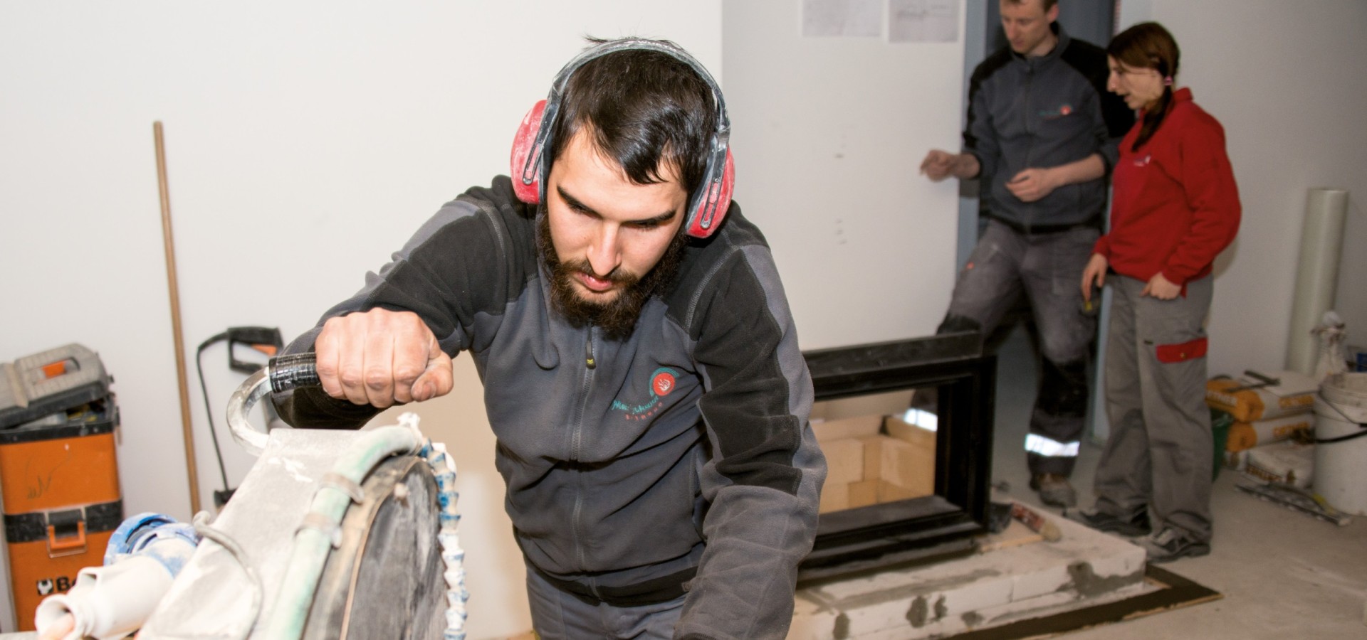 Person mit Gehörschutz bedient eine große Kreissäge in einer Werkstatt, im Hintergrund zwei weitere Personen