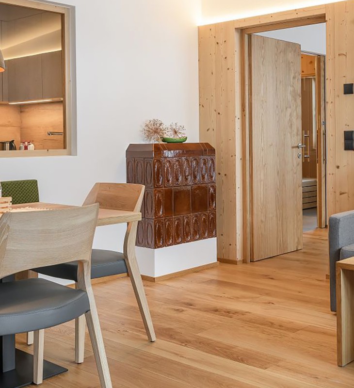 Essbereich mit Holztisch, Stühlen und grüner Polsterbank neben einem braunen Kachelofen in einem Raum mit Holzboden und offener Tür zu einem Flur