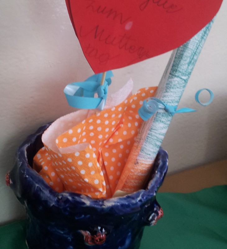 Blauer Keramiktopf mit orange-weiß gepunktetem Papier, einem gerollten Papierstreifen und einem roten Herz aus Papier mit handgeschriebenem Text