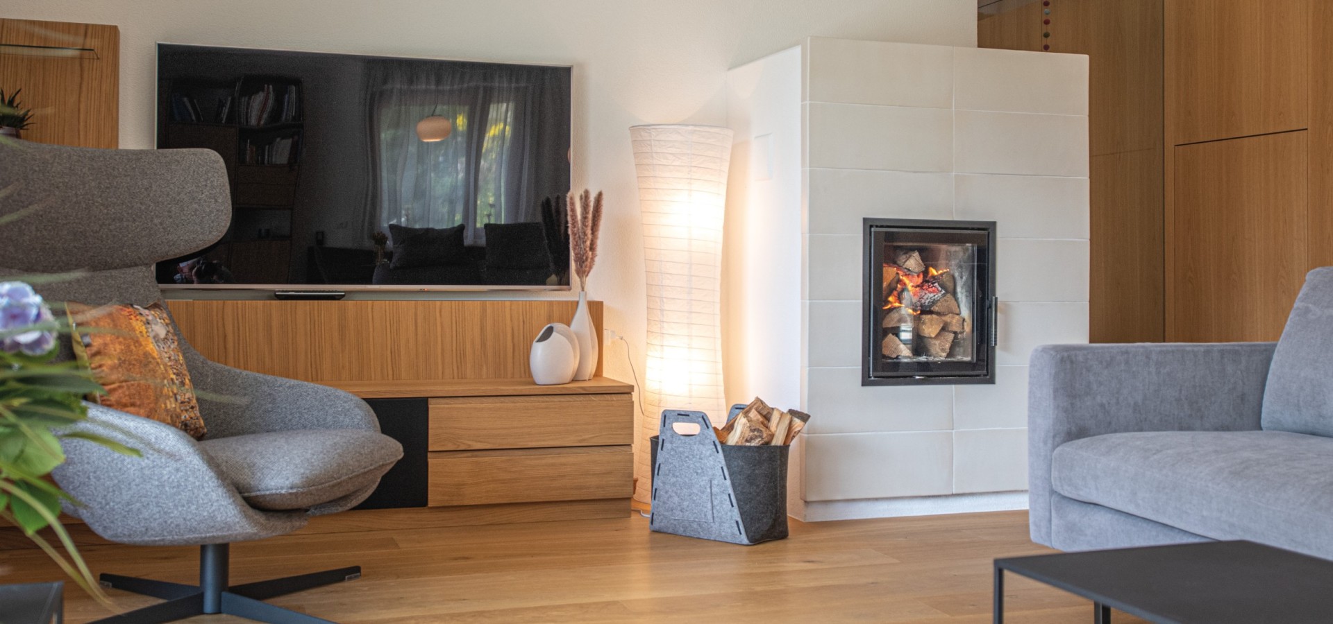 Wohnzimmer mit grauem Sessel, grauem Sofa, Holzboden, moderner weißer Kachelofen mit Feuer, Holzregal und Stehlampe