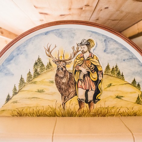 Halbrundes Bild mit einem Mann in historischer Kleidung neben einem Hirsch vor einer Landschaft mit Bäumen auf einem Bauernofen
