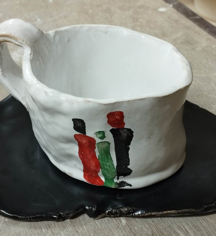 Unregelmäßig geformte weiße Keramiktasse mit unregelmäßigem Henkel und schwarzer, unregelmäßig geformter Untertasse mit roten, grünen und schwarzen Farbflecken auf der Tasse