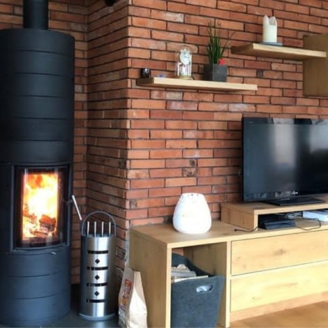 Schwarzer runder Kaminofen mit brennendem Feuer vor roter Ziegelwand neben Holzregalen mit Fernseher und Dekoration