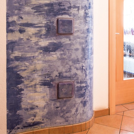 Runde Ofenwand mit strukturierter, blau-grauer Oberfläche und zwei quadratischen, braunen Steckdosen neben einer Holztür mit Glasfenstern und Fliesenboden
