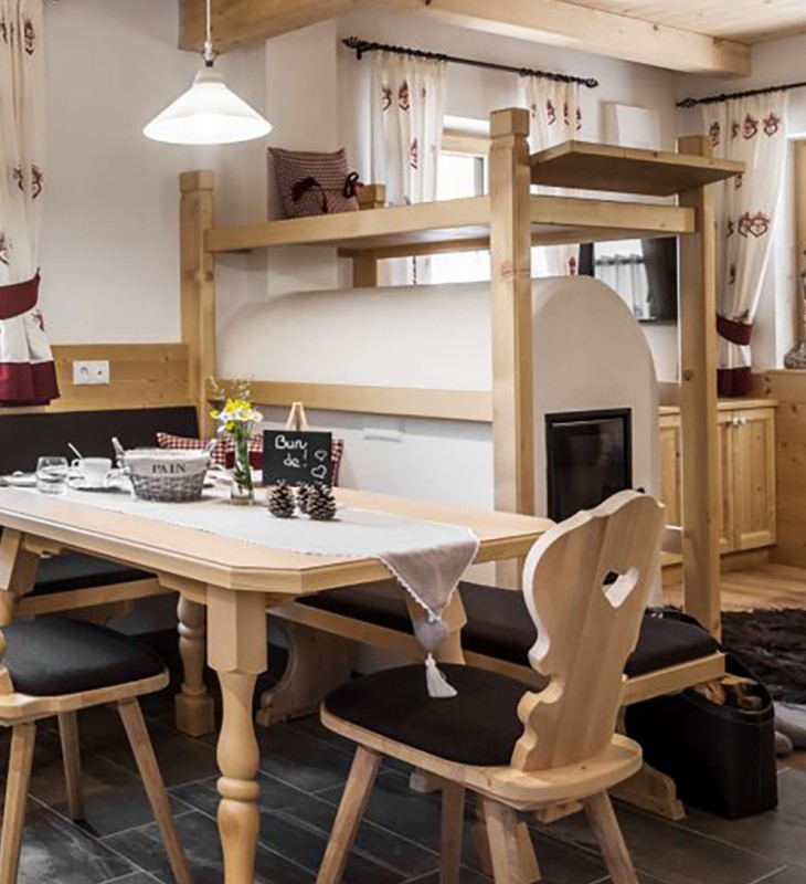 Ein gedeckter Holztisch mit zwei Holzstühlen, ein rustikaler Ofen mit Eckbank in einem gemütlichen, holzgetäfelten Zimmer