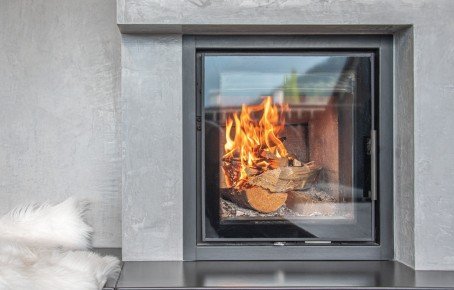 Moderner Kamin mit brennendem Holz in einem grauen Betonrahmen neben einem weißen Fellkissen