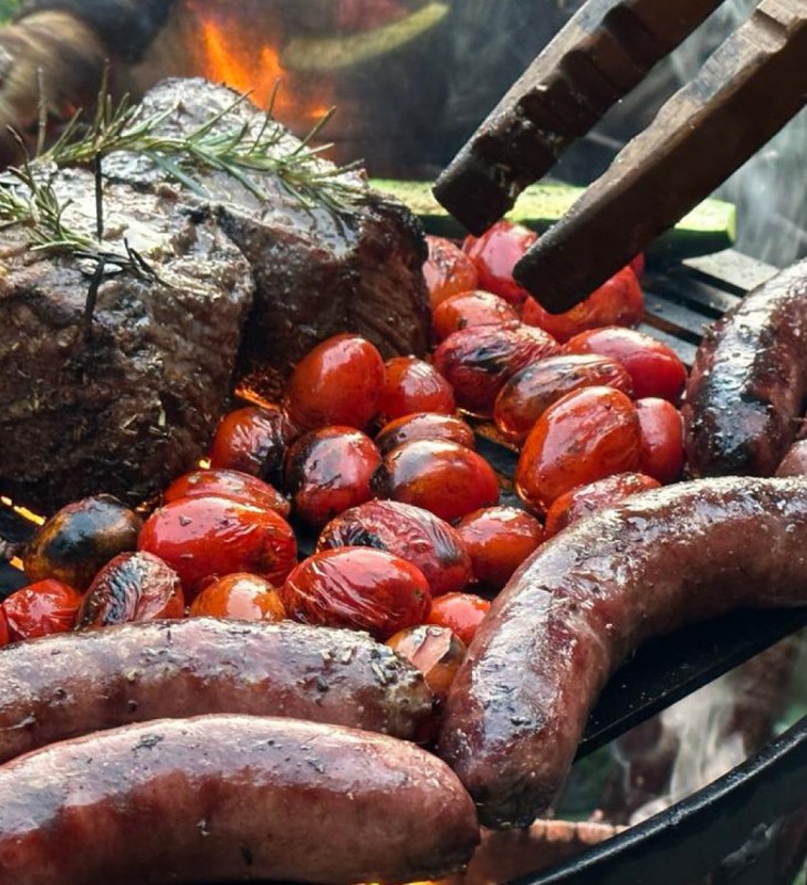 Gegrillte Würste, Tomaten und ein Stück Fleisch mit Rosmarin auf einem Grillrost über offenem Feuer