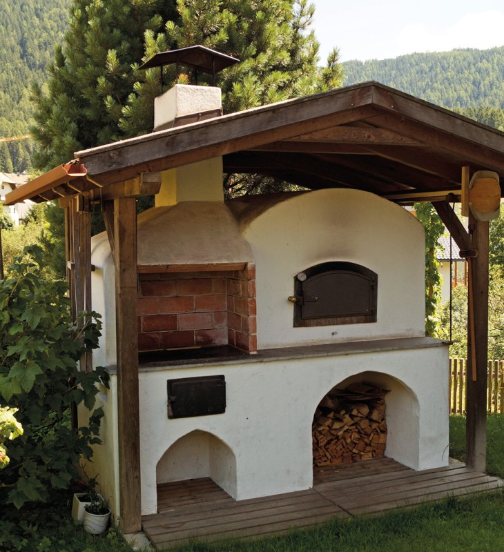 Freistehender, weiß verputzter Holzbackofen mit Schornstein und Holzlager unter einem Holzdach in einer grünen Berglandschaft