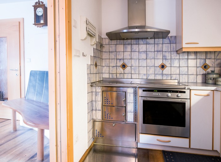 Kleine Küche mit Edelstahlherd, Backofen, Dunstabzugshaube und gefliester Wand neben einem offenen Durchgang zu einem Raum mit Holztisch und Tür