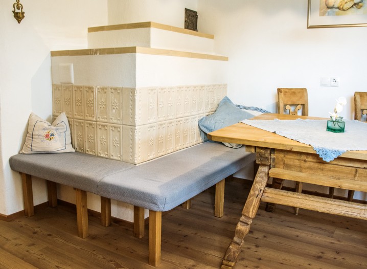 Eckbank mit gepolstertem Sitz und weißem Kachelofen mit goldfarbenen Zierstreifen in einem Raum mit Holzboden und Holztisch