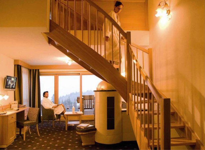 Hotelzimmer mit Teppichboden, einem modernen runden Kamin, Holztreppe und zwei Personen in Bademänteln, eine sitzt am Fenster, die andere steht auf der Treppe