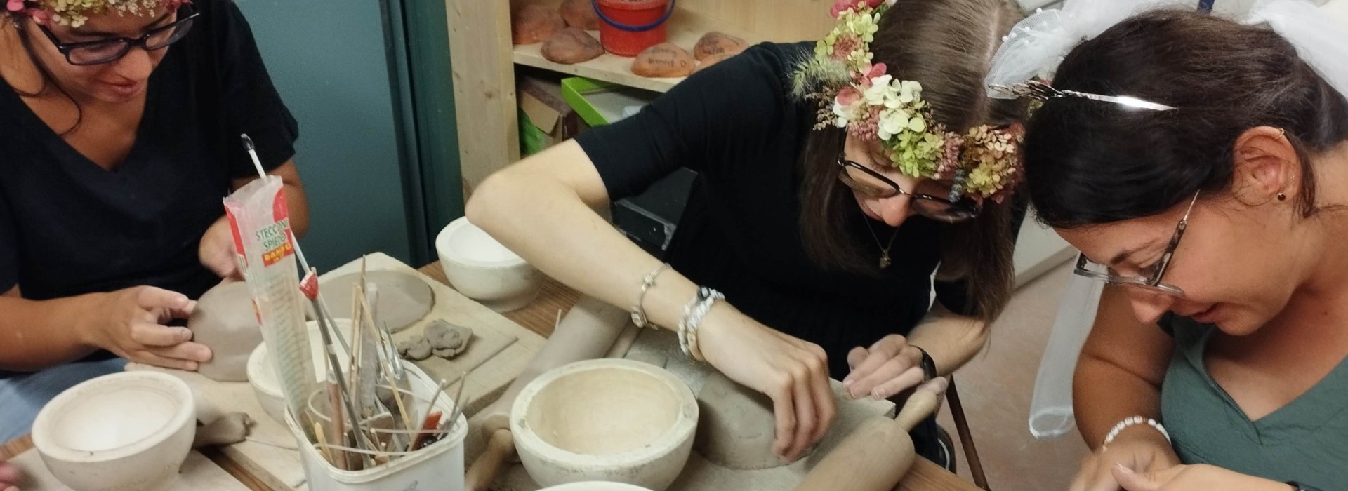 Drei Personen mit Blumenkränzen arbeiten an einem Tisch mit Töpferwerkzeugen und Ton