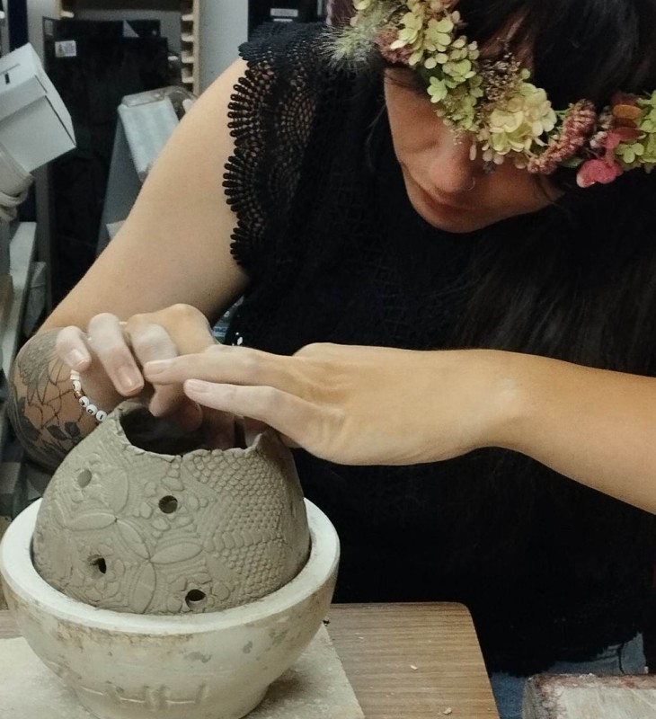 Frau mit Blumenkranz auf dem Kopf arbeitet an einem Tonobjekt mit Mustern und Löchern