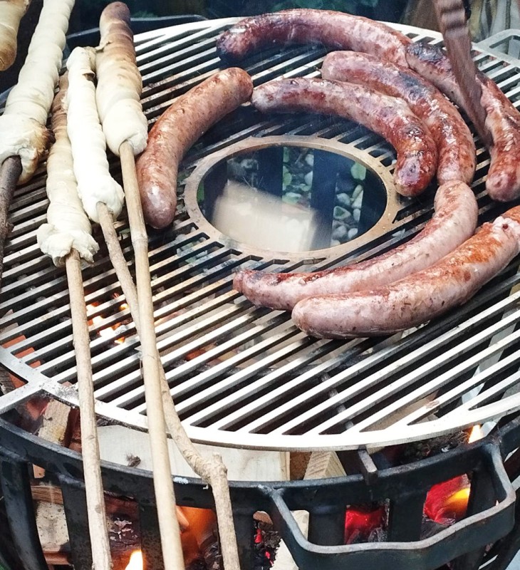 Mehrere Würstchen und mit Teig umwickelte Stangen liegen auf einem runden Grillrost über einer offenen Flamme