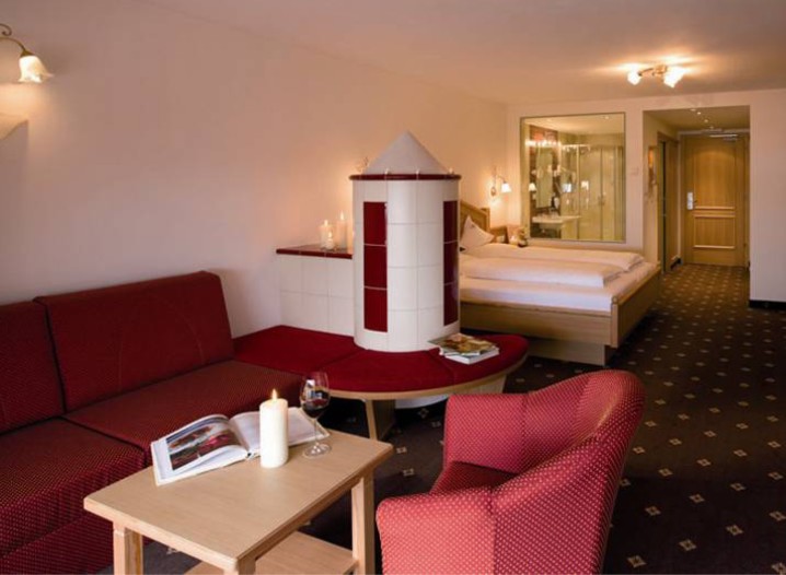 Hotelzimmer mit rotem Sofa, kleinem Tisch mit Kerze und Glas, rotem Sessel, rundem Kamin und Doppelbett im Hintergrund