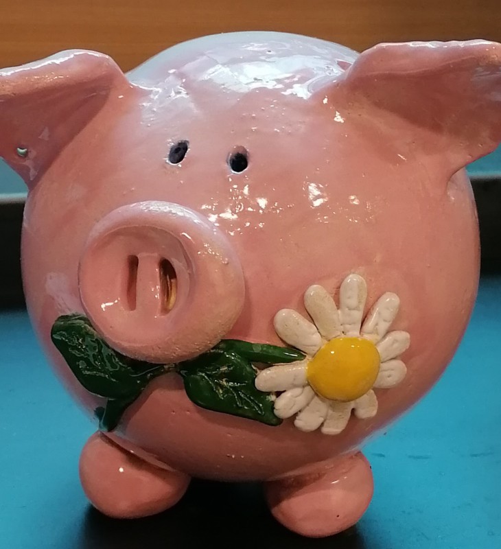 Rosa Sparschwein in Keramik mit aufgesetzter weiß-gelber Blume und grünen Blättern auf blauem Untergrund