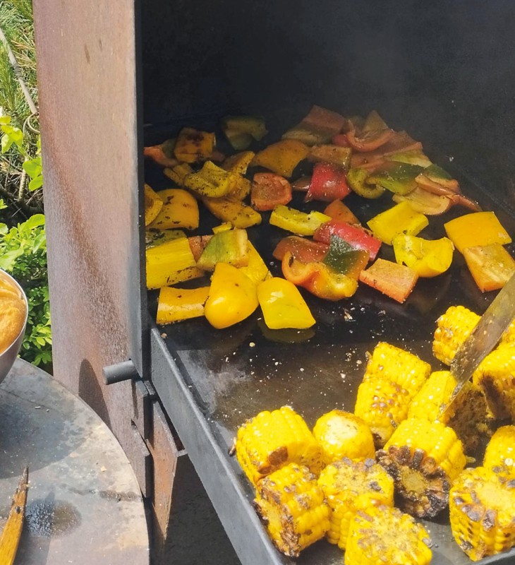 Gegrillte Maiskolben und Paprikastücke auf einer Grillplatte neben einer Schale mit Brötchen und einer Grillzange