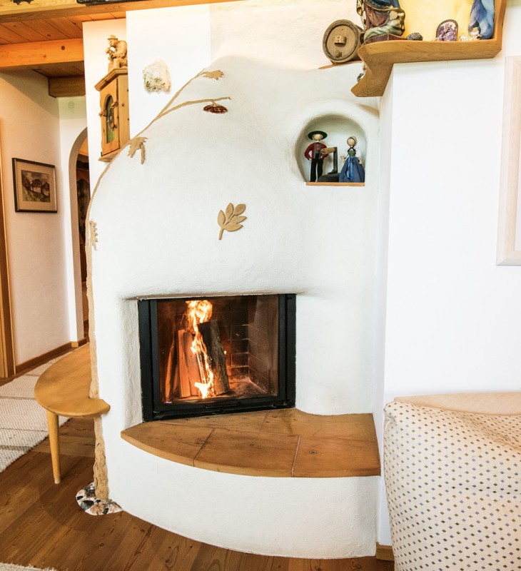 Wohnzimmer mit weißem Kachelofen mit Holzfeuer, Holzboden, Holzbalkendecke und offener Durchgang zur Küche