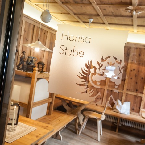 Holzgastraum mit Tischen und Stühlen hinter einer Glasscheibe mit der Aufschrift Honsa Stube und einem Wappensymbol