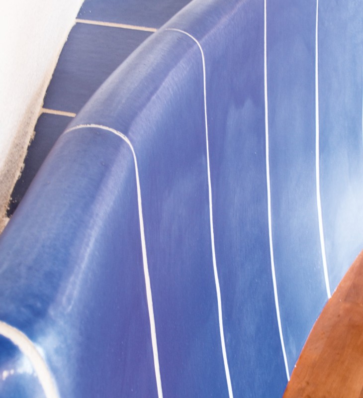 Blau gekachelte, gebogene Sitzbank mit weißen Linien. Die Oberfläche wirkt glänzend und glatt. Rechts im Bild ist eine hölzerne Tischplatte sichtbar. Die Wand links ist weiß und rau