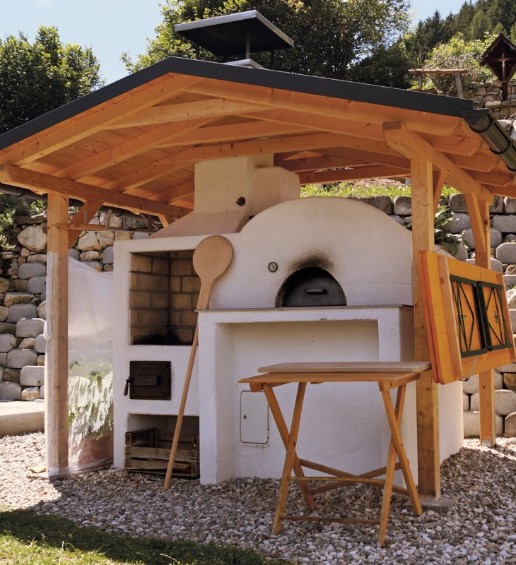 Freistehender Holzbackofen mit Dach, Holzlöffel und Klapptisch in einem Garten mit Steinmauer im Hintergrund