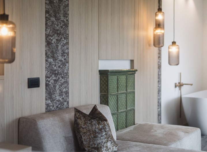 Wohnzimmer mit grauem Sofa, gemustertem Kissen, grünem Kachelofen und drei hängenden Lampen vor holzvertäfelter Wand