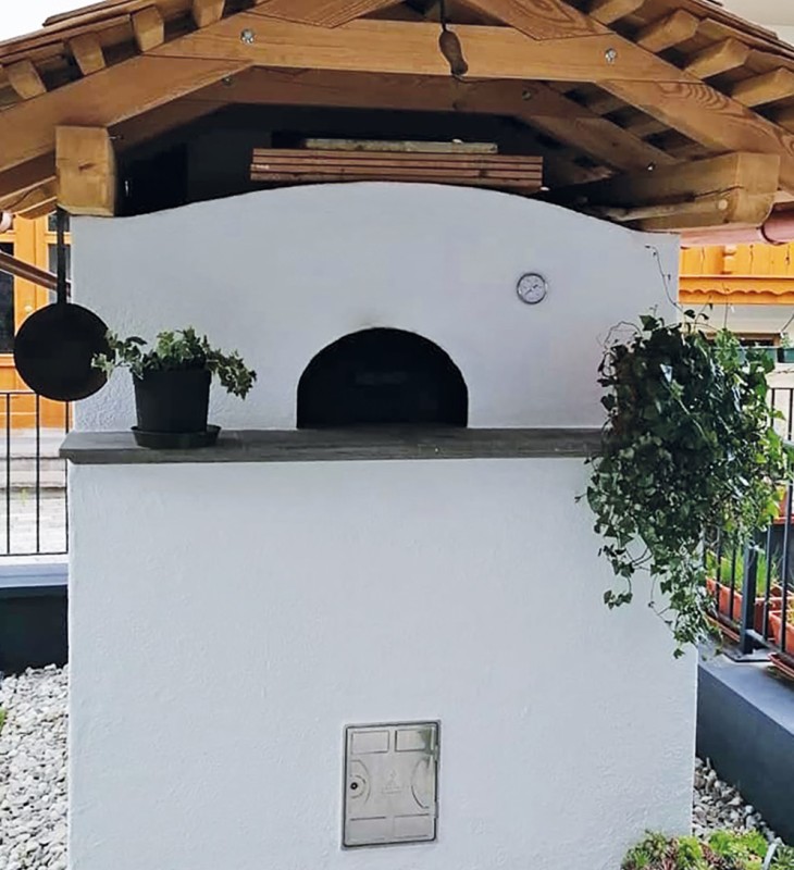 Weißer, gemauerter Außenofen mit rundem Backraum unter einem Holzdach, umgeben von Pflanzen und Gartenzaun