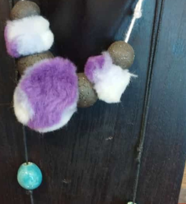 Schmuckstück mit lila-weißen und braunen Filzkugeln sowie einer blauen Perle vor dunklem Holz