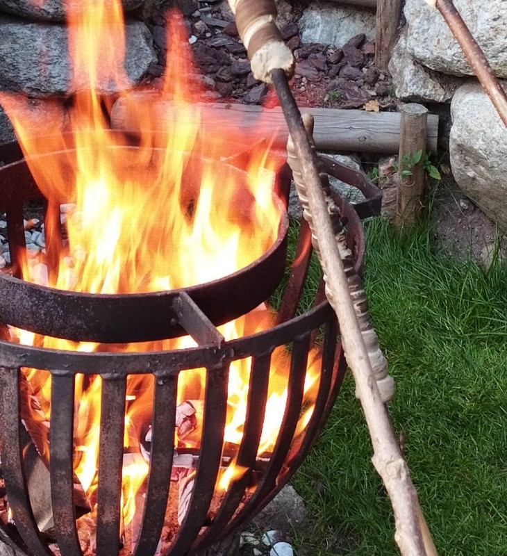 Feuerkorb mit brennendem Holz und zwei langen Stöcken, einer mit einer Wurst, auf Gras vor einer Steinmauer