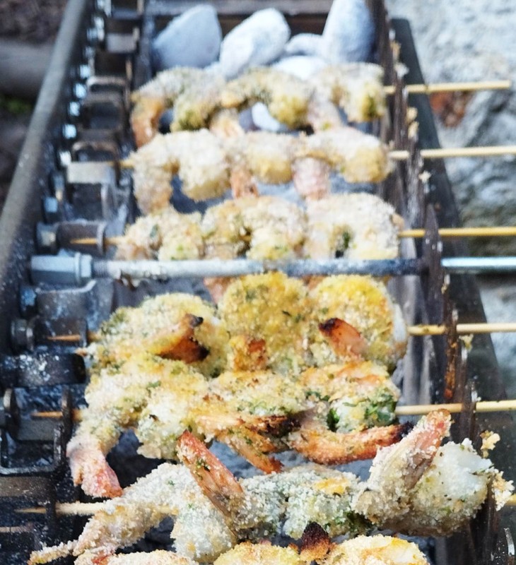 Mehrere Spieße mit panierten Garnelen liegen auf einem Grillrost über Holzkohle