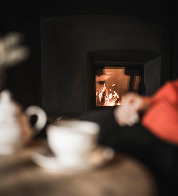 Unscharfer Vordergrund mit Teekanne und Tasse vor einem Kamin mit Feuer und einer Person in rotem Pullover