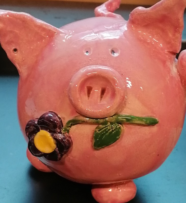 Rosa Sparschwein in Keramik mit aufgesetzter weiß-gelber Blume und grünen Blättern auf blauem Untergrund