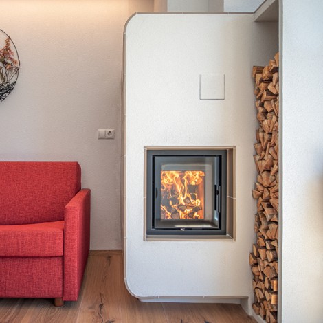 Moderner Kamin mit sichtbarem Feuer neben einem roten Sofa und einem Holzstapel in einem hellen Raum