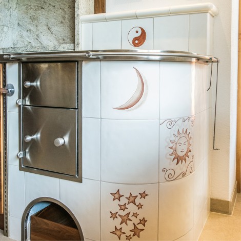 Weißer Kachelofen mit braunen Motiven von Mond, Sonne, Sternen und Yin-Yang-Symbol neben Holzküchenschränken