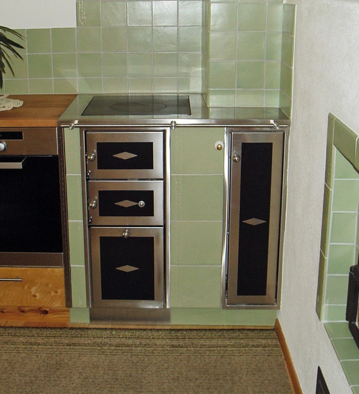 Kleine Küche mit grün gekachelter Wand, Holzarbeitsplatte, schwarzem Backofen und grün-schwarzem Holzofen mit Metallrahmen